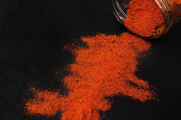 A vibrant mound of bright red-orange paprika powder spilled from a glass jar onto a dark, textured black background. High quality photo